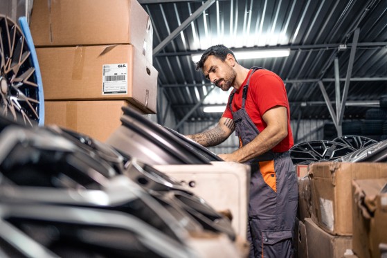 A warehouse worker inspects automotive parts beside stacked shipping boxes.